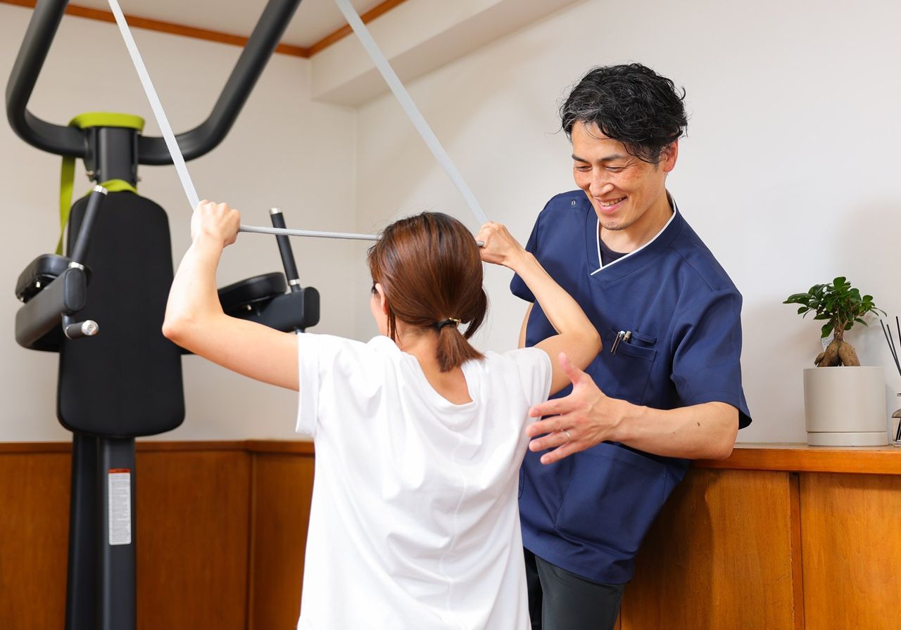 トレーニングマシンの前でゴムバンドを持つ利用者に対して、トレーナーが笑顔で肩まわりの動きを確認している場面